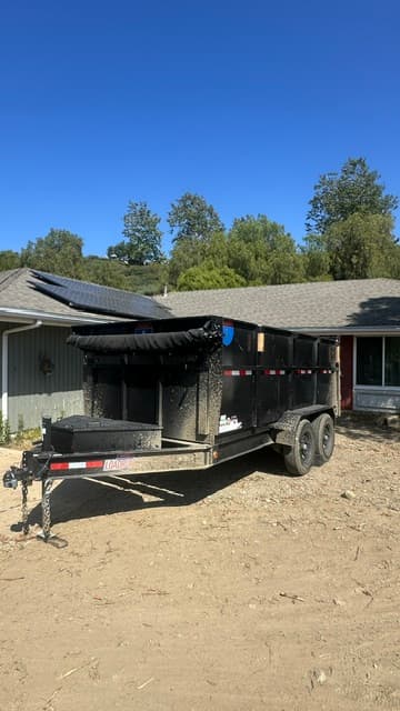 Black dual-axle dump trailer parked on a dirt lot in front of a residential house.