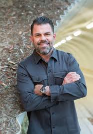 Smiling bearded man in a grey shirt poses with arms crossed in an outdoor setting.