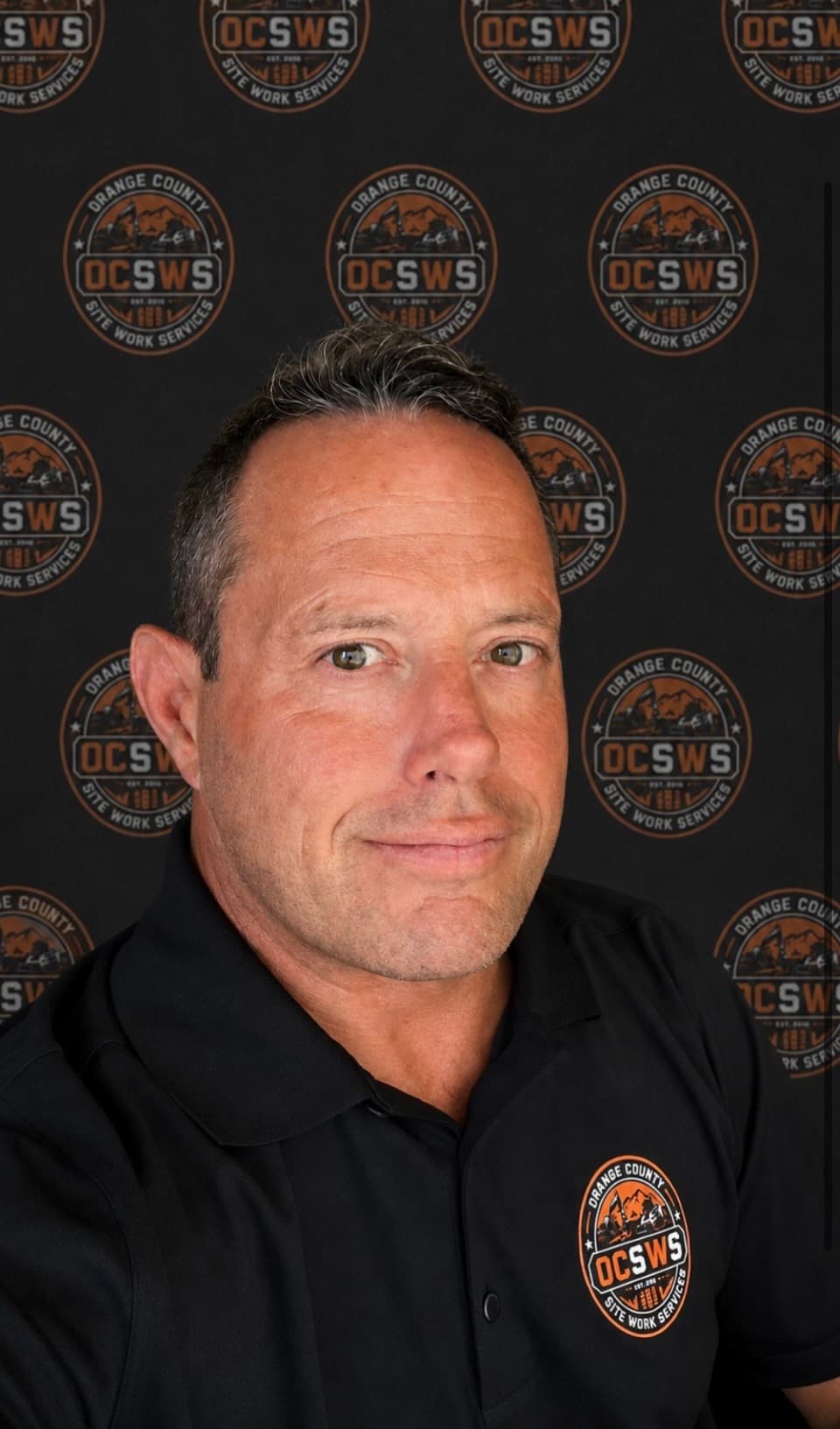 Man in black polo shirt smiling against a repeating Orange County OCSWS logo backdrop.